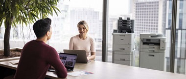 Zwei Büroangestellte arbeiten in einem hellen Büro an einem Schreibtisch. Im Hintergrund stehen zwei Brother KMU-Druckern vor einer städtischen 