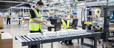 Eine Mitarbeiterin klebt an einem Förderband in einem Produktions- und Logistikbetrieb eine gedruckte Barcode-Etikette auf ein Paket.