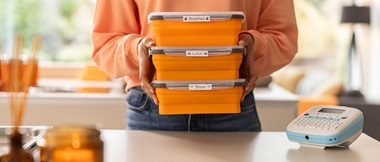 Woman holding 3 orange boxes with labels stacked on top of each other in front of a table with a Brother Btag label printer