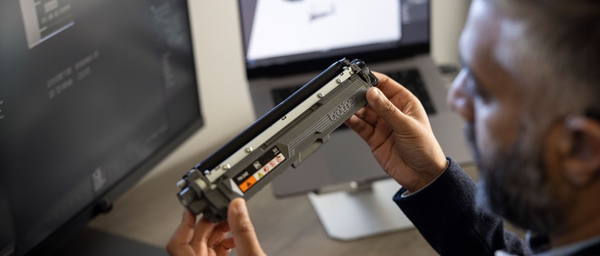 Man holding up a Brother toner cartridge with two hands to inspect it with computer screens in the background