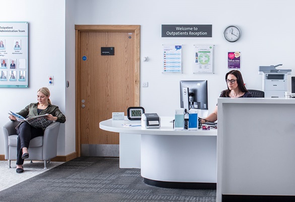Female receptionist sat at desk using a computer, female patient sat reading newspaper
