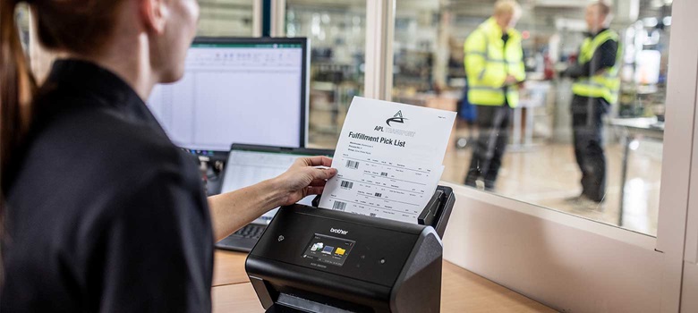 Woman in black holding document in scanner, warehouse, laptop, computer screen, people wearing hi-vis in warehouse